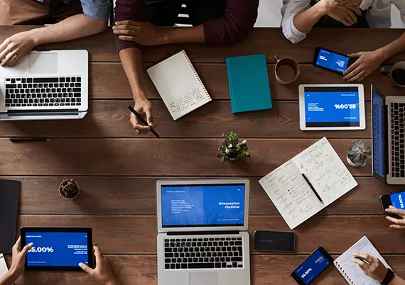top view of people near wooden table having multiple gadgets
              on it like laptop, mobile, tablets